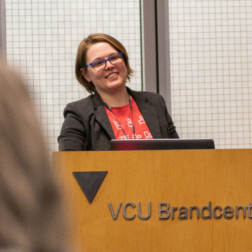 Portrait of Caitlyn standing at a podium in VCU Brandcenter, brown hair and blue framed glasses wearing a grey blazer over a red tshirt smiling towards the audience.