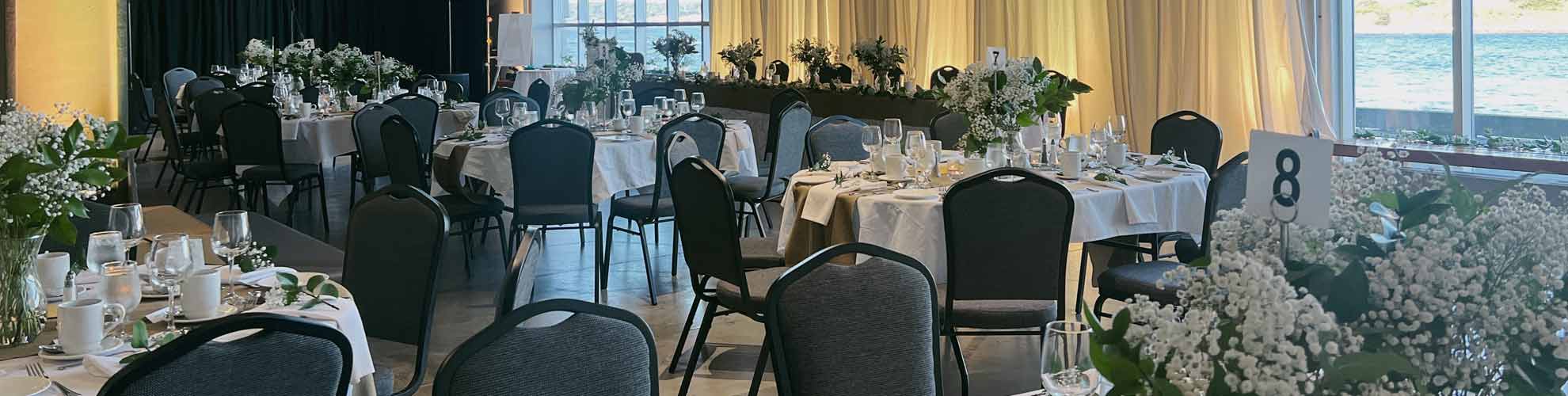 Wedding banquet set with decorated oval guest tables and a long head table, a small stage for a band, and large floor to ceiling windows. 