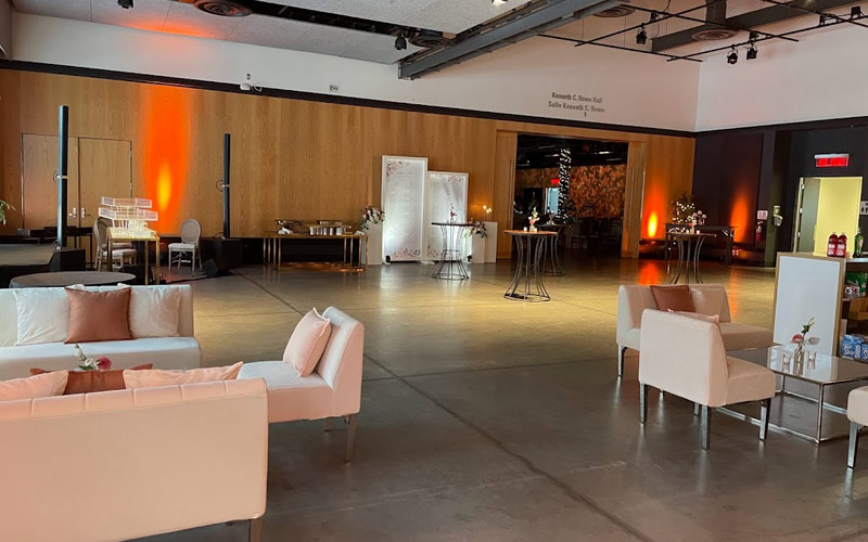 The room is set for a reception with white club chairs and sofas around two glass top coffee tables and tall decorated cruisers tables. A large seating chart is displayed on a set of white frames.