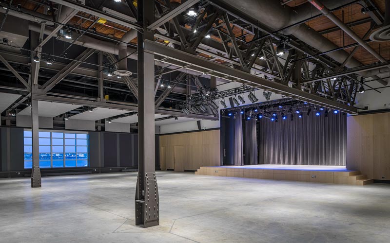 An empty venue space with a tall ceiling, large stage with professional lighting, grey velour curtains, polished concrete flooring, a wall installed with sound panels, and a large window that provides a view of the Halifax Harbour.