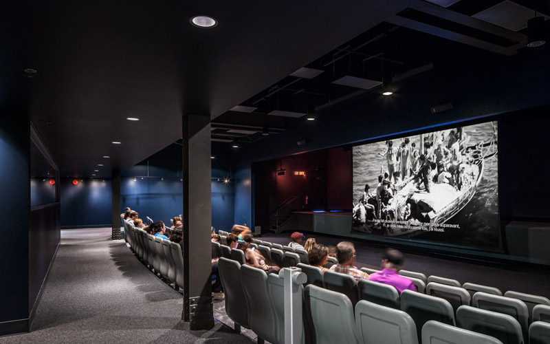 An amphitheater with one hundred chairs, and a dozen guests sitting and facing a drop-down screen with a projected black and white image of a ship.