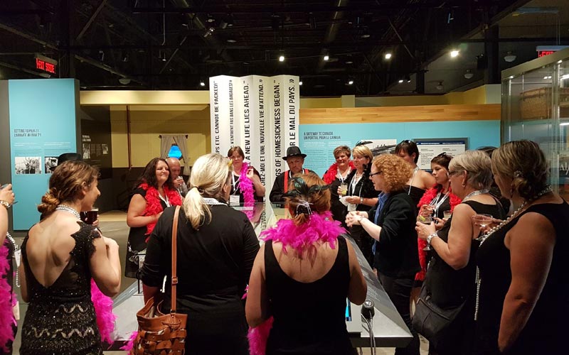 A group of visitors dressed in cocktail attire and feather boas stand around an interpreter overlooking a model of the historic site. 