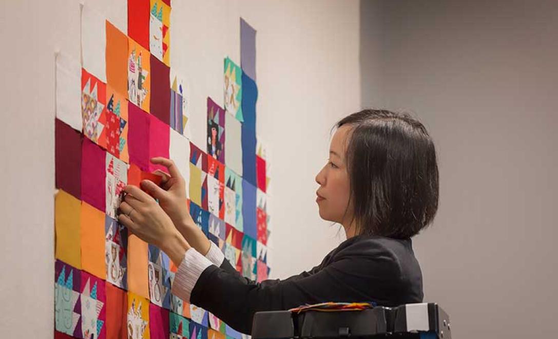 A woman works on a quilt that is hanging on a wall.