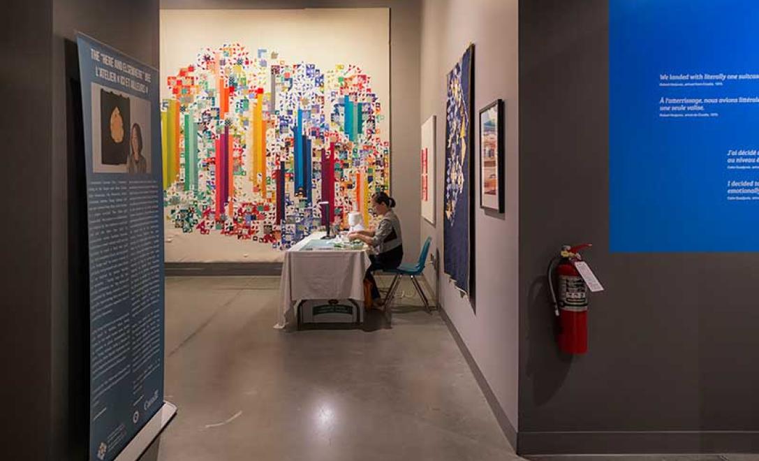 Quilt and woman working on a sewing machine can be seen from a distance.