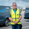 Juan Antonio Carrasco, reconocido como Driver Hero. Está posando mirando a cámara, con las manos cruzadas delane y de fondo furgonetas de color gris con el logo, sonrisa de Amazon, en azul y la palabra Prime en blanco. Él está con las manos cruzadas delante, un polo azul de manga corta y una chaleco de color amarillo. Tiene el pelo corto negro y blanco. 