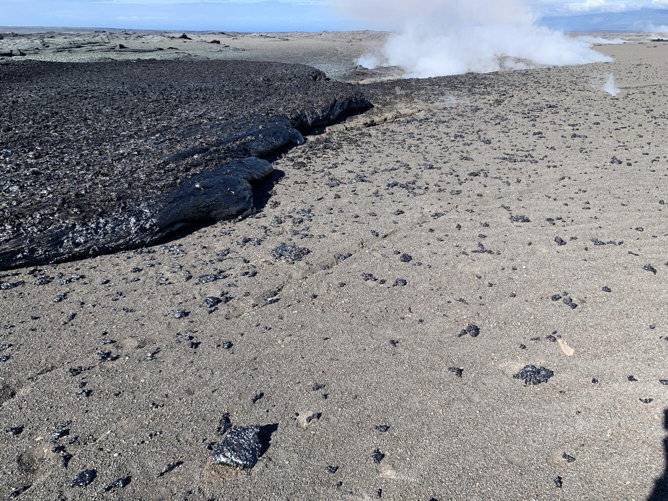 A shiny black lava flow is bordered by angular, frothy fragments of glassy rock scattered over a gravelly landscape