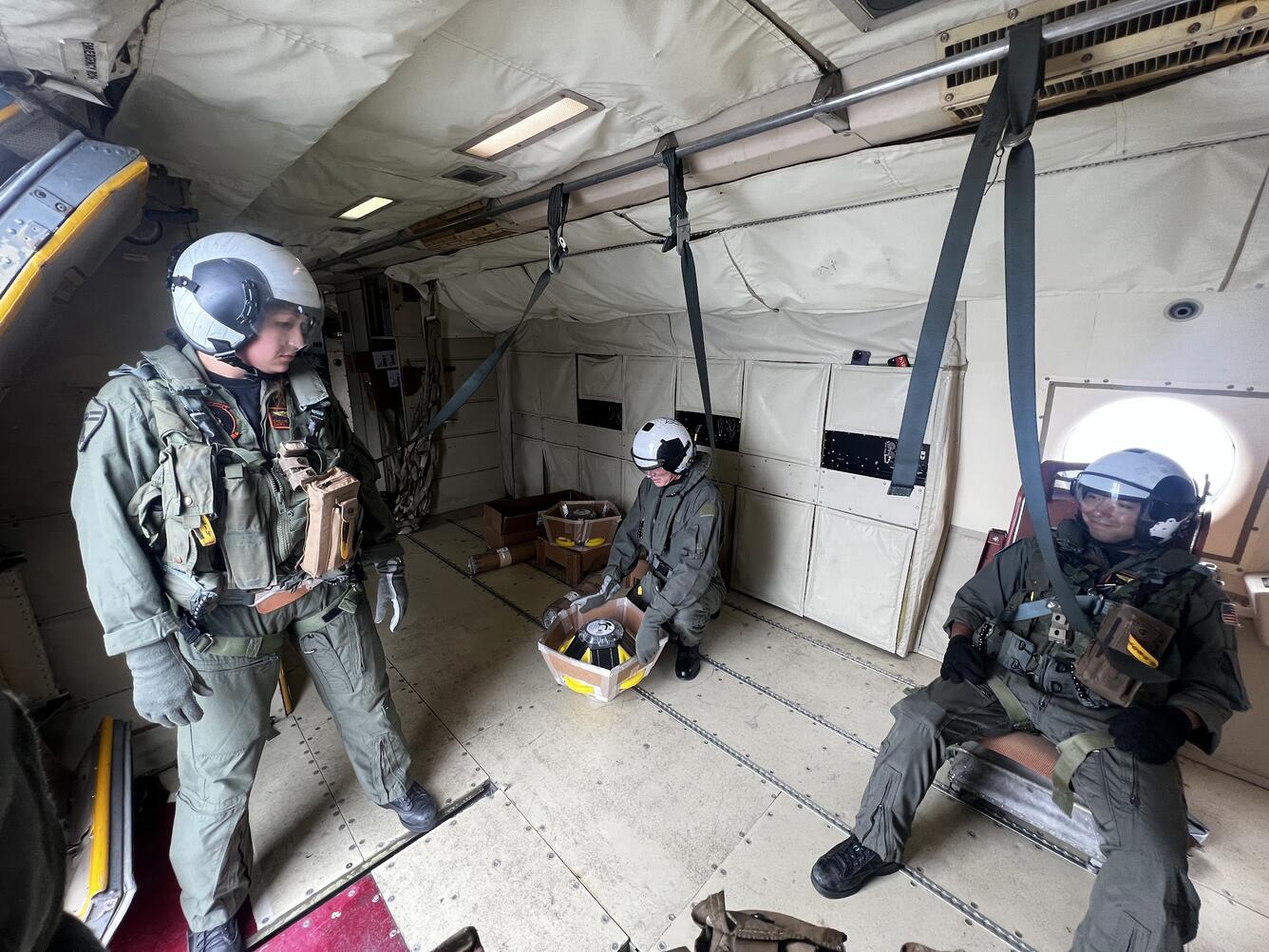 Three people inside a plane wearing flight suites and helmets, two are seated, one is standing looking at them