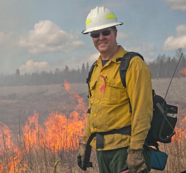 A man wearing protective gear stands in front of a low prescribed fire in the background