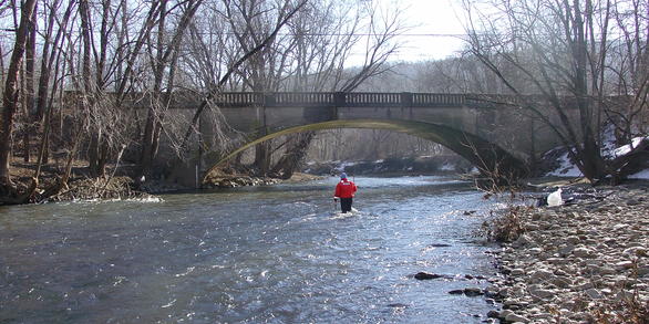 USGS Technician wading across a river toward an arch bridge.