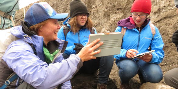 Three girls sit on rocks and look at a computer screen.