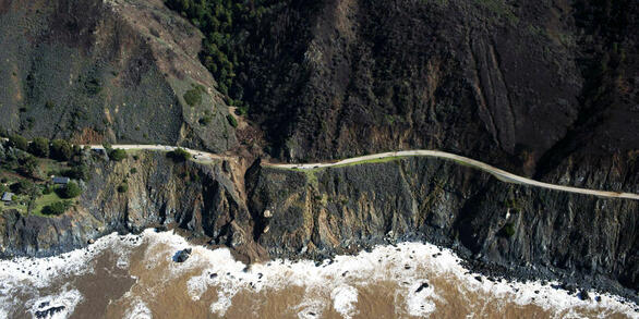 A view from the sky of a winding coastal road that runs along steep, rocky cliffs, with a section washed out and collapsed.