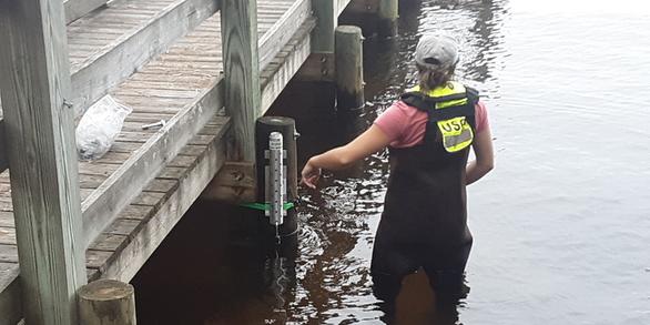  A USGS employee standing in knee deep water attaches a metal cylinder containing a storm tide sensor to a pier. 