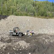 Two scientists next to 4x4 vehicle and buckets. Gravel-rock hill, tire tracks in mud, green trees, blue sky in background.