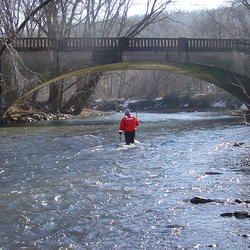 USGS Technician wading across a river toward an arch bridge.