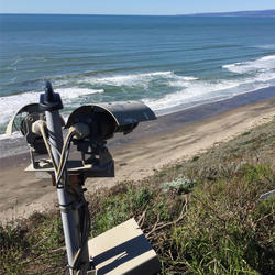 A pole has two shaded cameras mounted on it and they are pointing at a beach from high up on a grassy bluff.