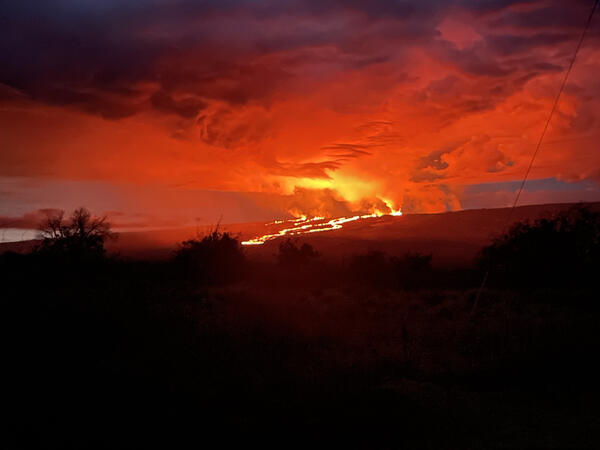 glowing red and orange lava flows moving down a dark slope with orange and red clouds in the sky.