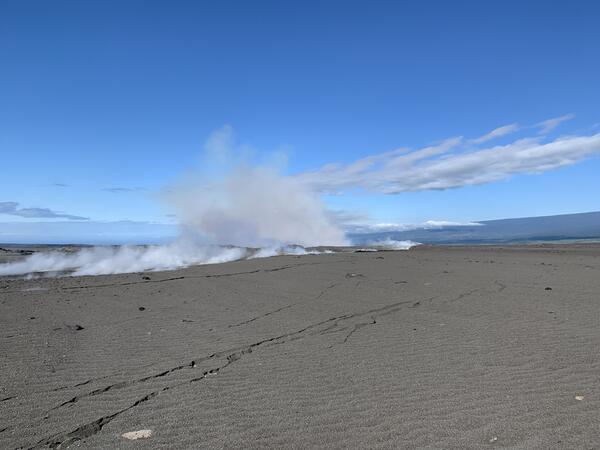 Ground cracks spider into the distance on a gravelly black landscape. In the background, bigger cracks emit plumes of gas