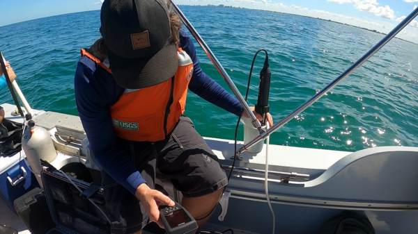 a scientist sits on the edge of a boat on the ocean with an instrument submerged in a sample tube of water