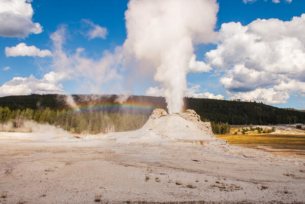 A stream of water and steam erupts from a geyser while a rainbow crosses over top.