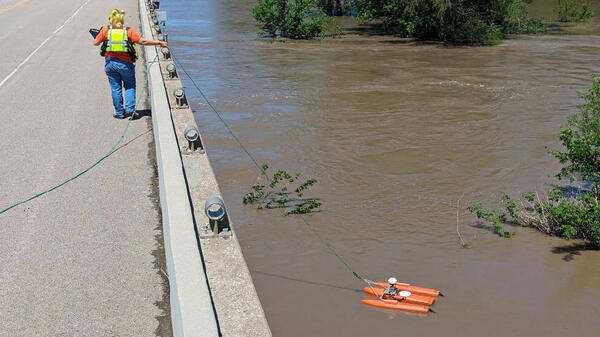 A USGS hydrologic technician on a bridge guides an orange streamflow measuring device across a river to measure flooding