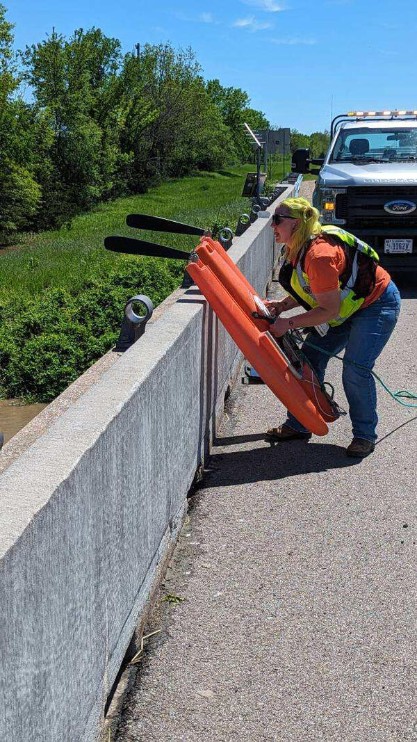 A USGS hydrologic technician prepares to lift an orange streamflow measuring device onto the ledge of a bridge.