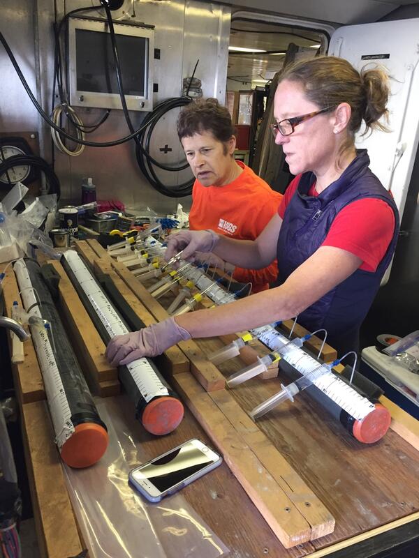 Two women stand at a table having a discussion while looking at long cores of sediment samples.