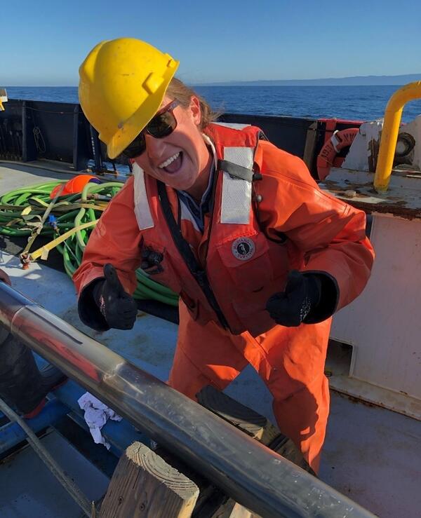 A smiling woman in a hard hat and life jacket gives two-thumbs-up on the deck of a ship standing next to a sediment core tube.