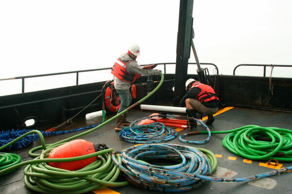 Two men work on a ship's deck with piles of cables, surrounded by thick fog