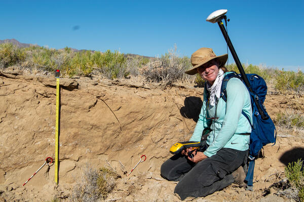 a woman with a GPS antenna kneels along an exposed rock face that is being measured 