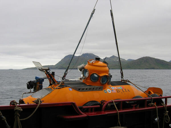 A submersible hanging from cables on the side of a ship in very calm water, with mountains far off in the distance.