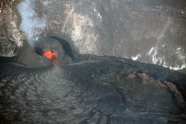 Color photo of a lava fountain within a black spatter cone at the edge of a lava lake