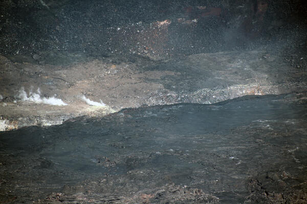 A telephoto color image of dark stagnant lava lake surface crust along the margin, with older crater wall at the edge