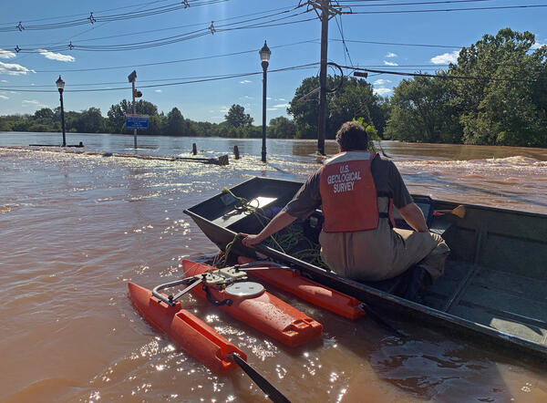 Image shows a USGS scientist in a PFD sitting in a boat with a red floating device alongside floating in floodwaters