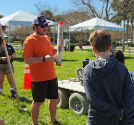 A USGS employee holds a display sediment core outside in front of a group of schoolchildren