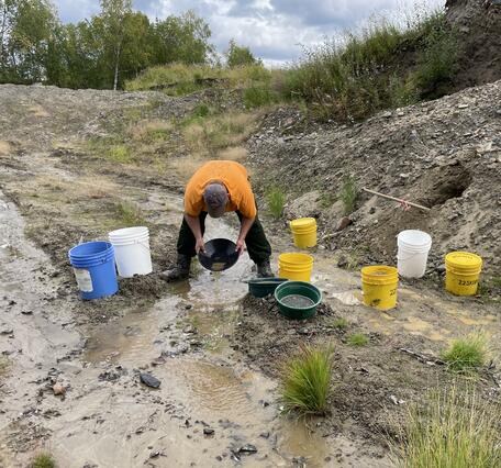 Scientist in orange shirt and hat with gold pan in hands. Seven buckets scattered around shallow creek and small hills.