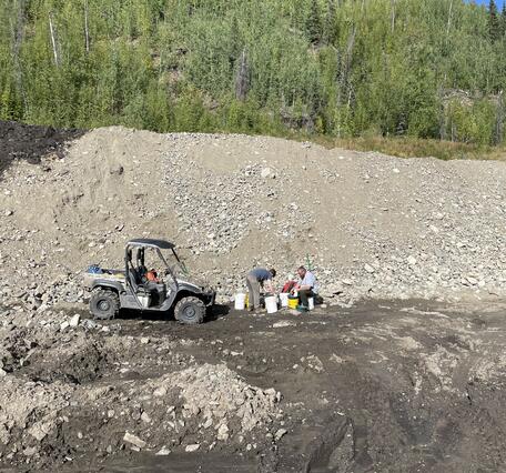 Two scientists next to 4x4 vehicle and buckets. Gravel-rock hill, tire tracks in mud, green trees, blue sky in background.