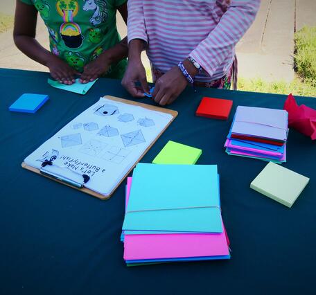 two sets of children's hands folding colorful paper on a table outside