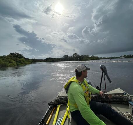 Man in a bright green shirt wearing a baseball cap guides a boat over water 