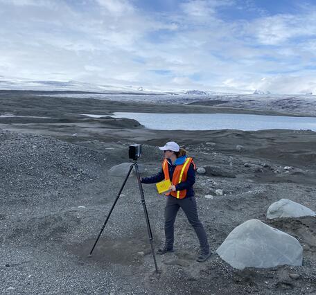 A scientist stands next to a tripod as she prepares to take measurements. Her surroundings are loose rocks and dirt.