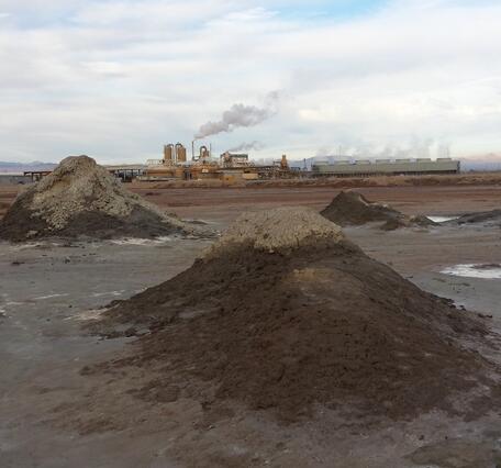 In the foreground, muddy mounds sit in the midst of pools of bubbling water. Behind, a geothermal power plant vents steam.