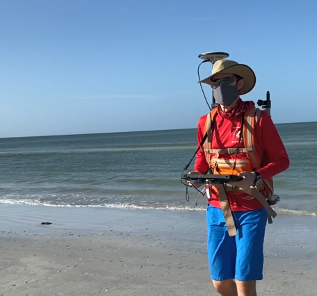 A man walks on a beach with a tablet in hand and a backpack on with scientific equipment extending from it. 