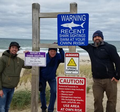 Photograph of USGS and University of KwaZulu-Natal personnel in front of a shark sign on the Lower/Outer Cape, Massachusetts