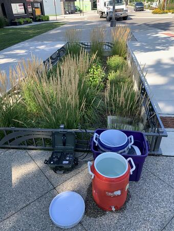 Two cylindrical coolers and water quality monitoring equipment next to greenery planted in a sidewalk urban environment.