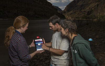 Three people gather around a glowing tablet along the Colorado River in Grand Canyon