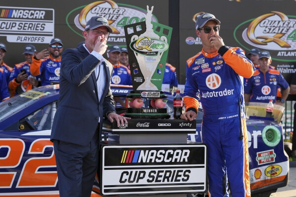 Joey Logano, right, is presented with the trophy and takes a traditional bite out of a peach in Victory Lane after winning a NASCAR Cup Series auto race, Sunday, Sept. 8, 2024, in Hampton, Ga. (AP Photo/Jason Allen)