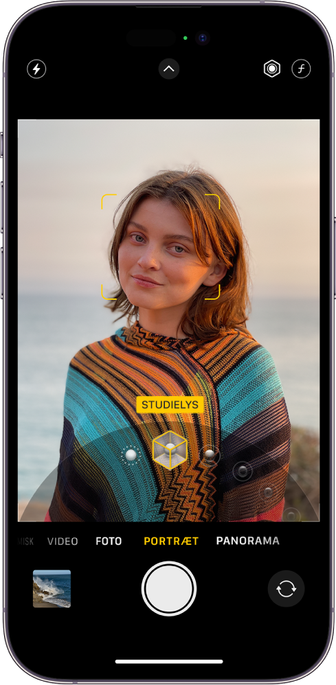 Kameraskærmen er i funktionen Portræt, i søgeren er motivet skarpt, og baggrunden er sløret. Drejeknappen til valg af portrætlys er åben nederst på billedet, og Studielys er valgt. Øverst til venstre på skærmen findes knappen Blitz, øverst i midten vises knappen Kameraindstillinger, og øverst til højre findes knapper til justering af intensiteten af portrætlys og Dybdekontrol. Nederst på skærmen fra venstre mod højre vises knappen Foto- og videofremviser, knappen Tag billede og knappen til valg af kamera på bagsiden.