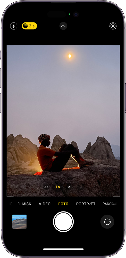 Kameraskærmen i funktionen Foto. I søgeren ses en natscene. Knapper til blitz og Nattilstand vises i øverste venstre hjørne af skærmen og Nattilstand er slået til. Knappen Kameraindstillinger findes øverst i midten, og knappen Live Photos findes i øverste højre hjørne. Nederst på skærmen fra venstre mod højre vises knappen Foto- og videofremviser, knappen Tag billede og knappen til valg af kamera på bagsiden.