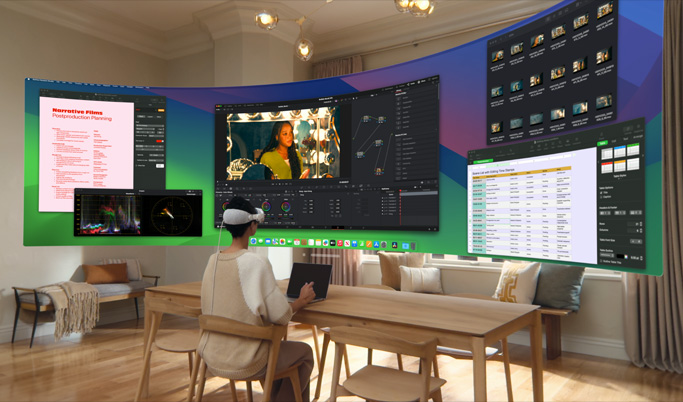 A woman sitting at a desk wearing Apple Vision Pro. In front of her, in 3D space, are mulitple screens each with a different app in use.