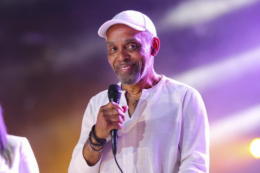 Frankie Beverly in white hat and linen shirt hold a microphone onstage