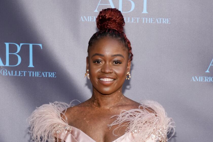 Michaela DePrince in a pale pink gown with feathers on the shoulders smiling at against a gray backdrop